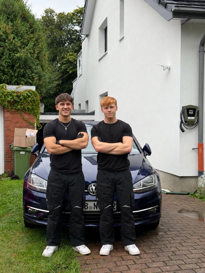 Eliah Stojanovic und David Göricke in schwarzer Arbeitskleidung vor einem dunkelblauen VW Golf
