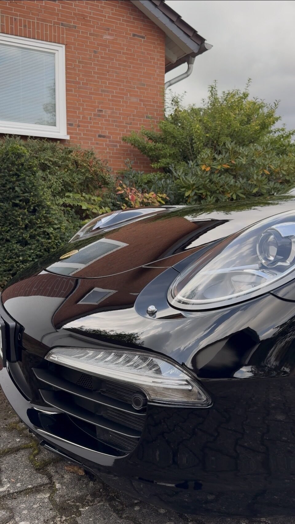 Front eines schwarzen Porsche nach der Aufbereitung: Haube, Kotflügel und Scheinwerfer spiegeln Hauswand, Büsche und Himmel klar
